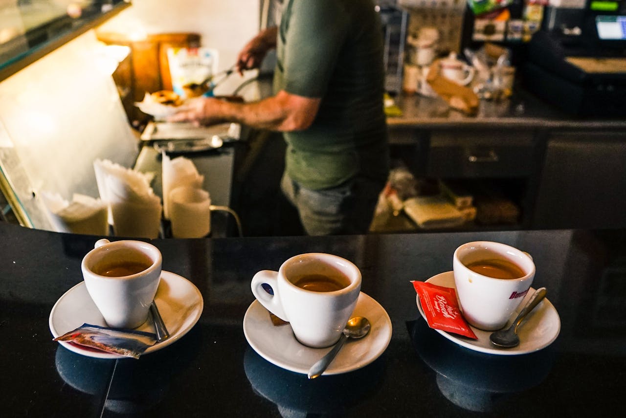 hero-img-02 A barista in a café preparing espresso drinks on a countertop with three coffee cups in focus.